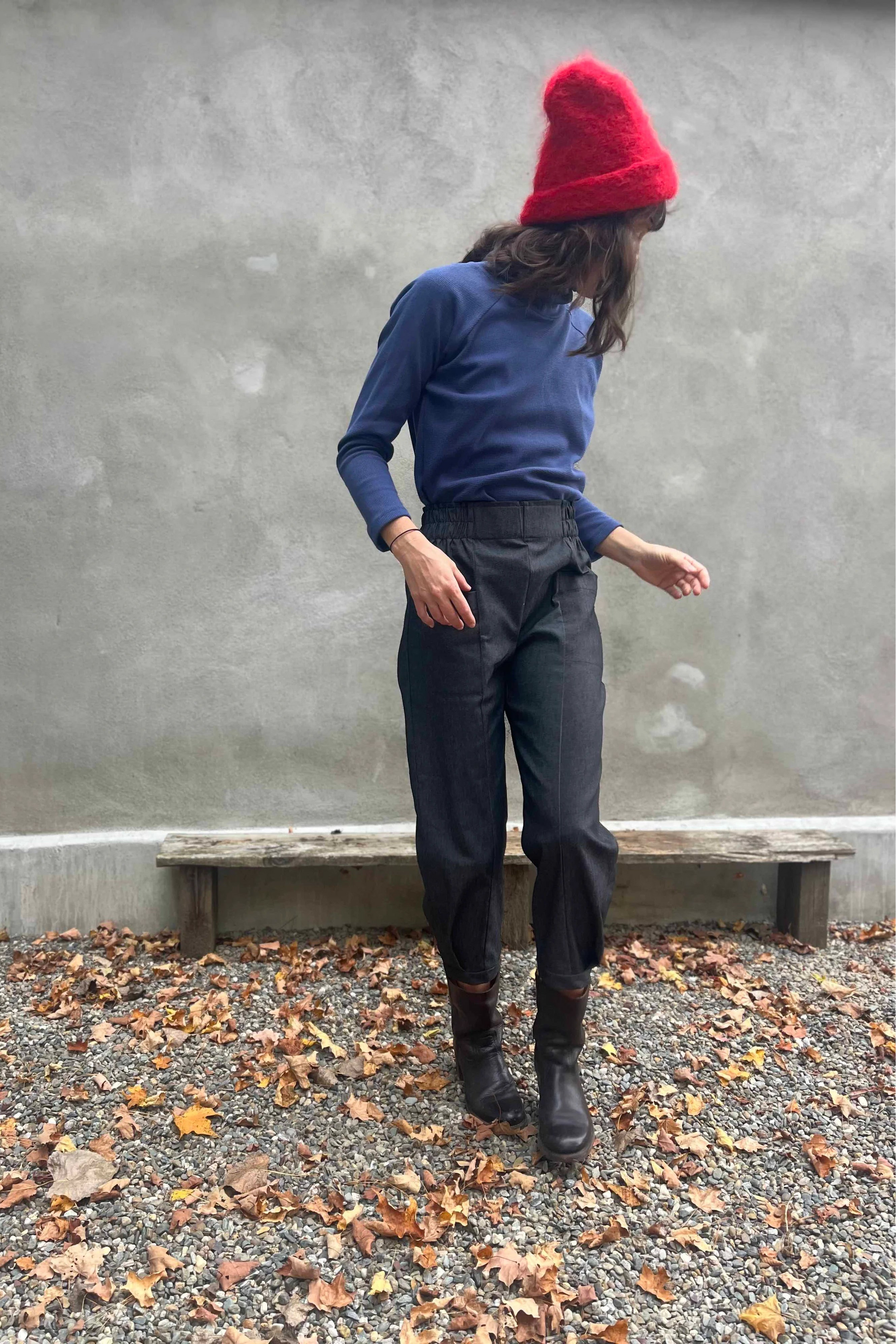 A woman wearing the Nyssa Pants by Kazak in Black Denim, barrel-legged pants with an elastic waistband and patch pockets. She is wearing them with the Oris Turtleneck in Indigo Waffle and standing outside by a grey wall. 