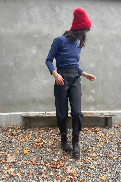 A woman wearing the Nyssa Pants by Kazak in Black Denim, barrel-legged pants with an elastic waistband and patch pockets. She is wearing them with the Oris Turtleneck in Indigo Waffle and standing outside by a grey wall. 