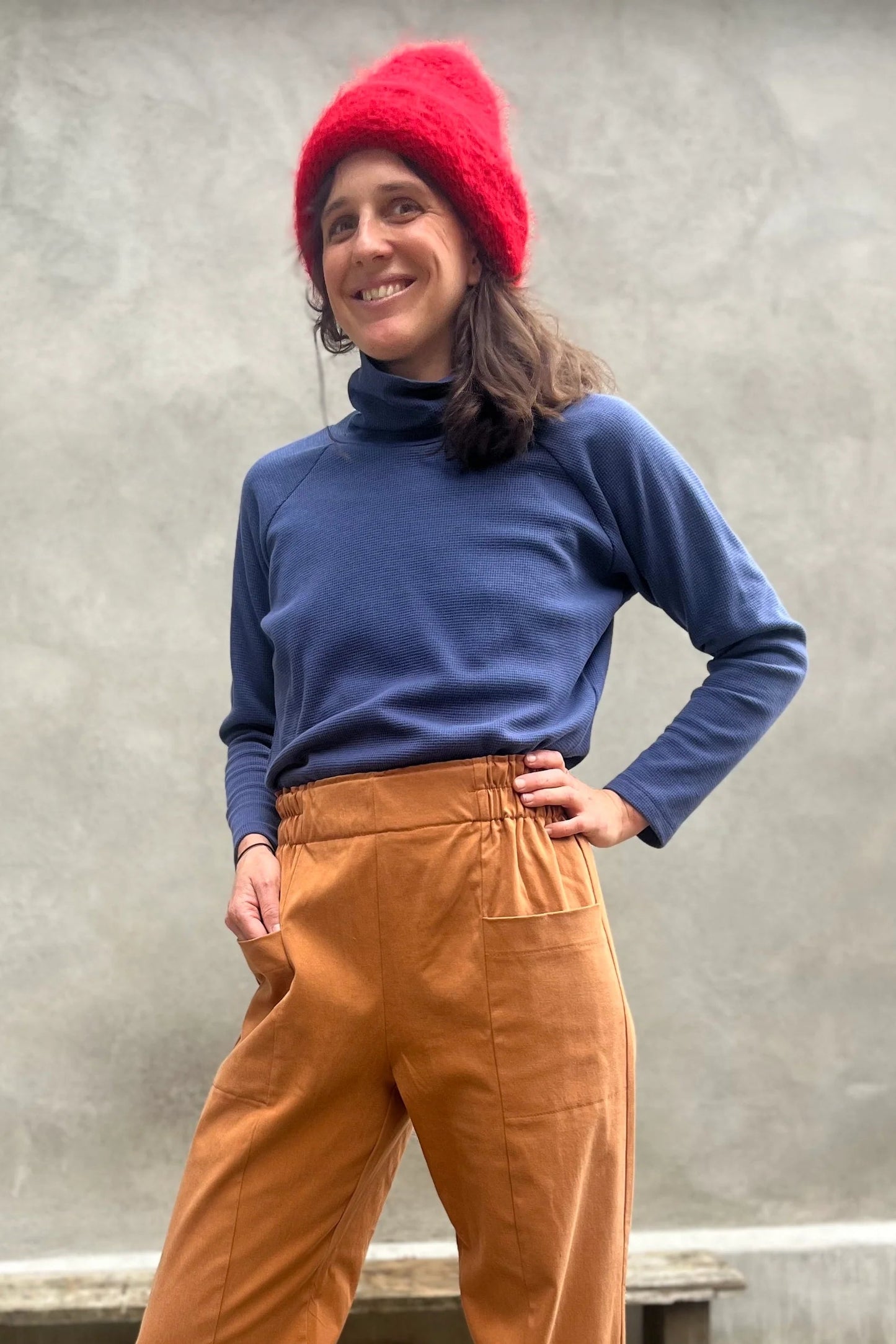A woman wearing the Nyssa Pants by Kazak in Terracotta, barrel-legged pants with an elastic waistband and patch pockets. She is wearing them with the Oris Turtleneck in Indigo Waffle and standing outside by a grey wall. 