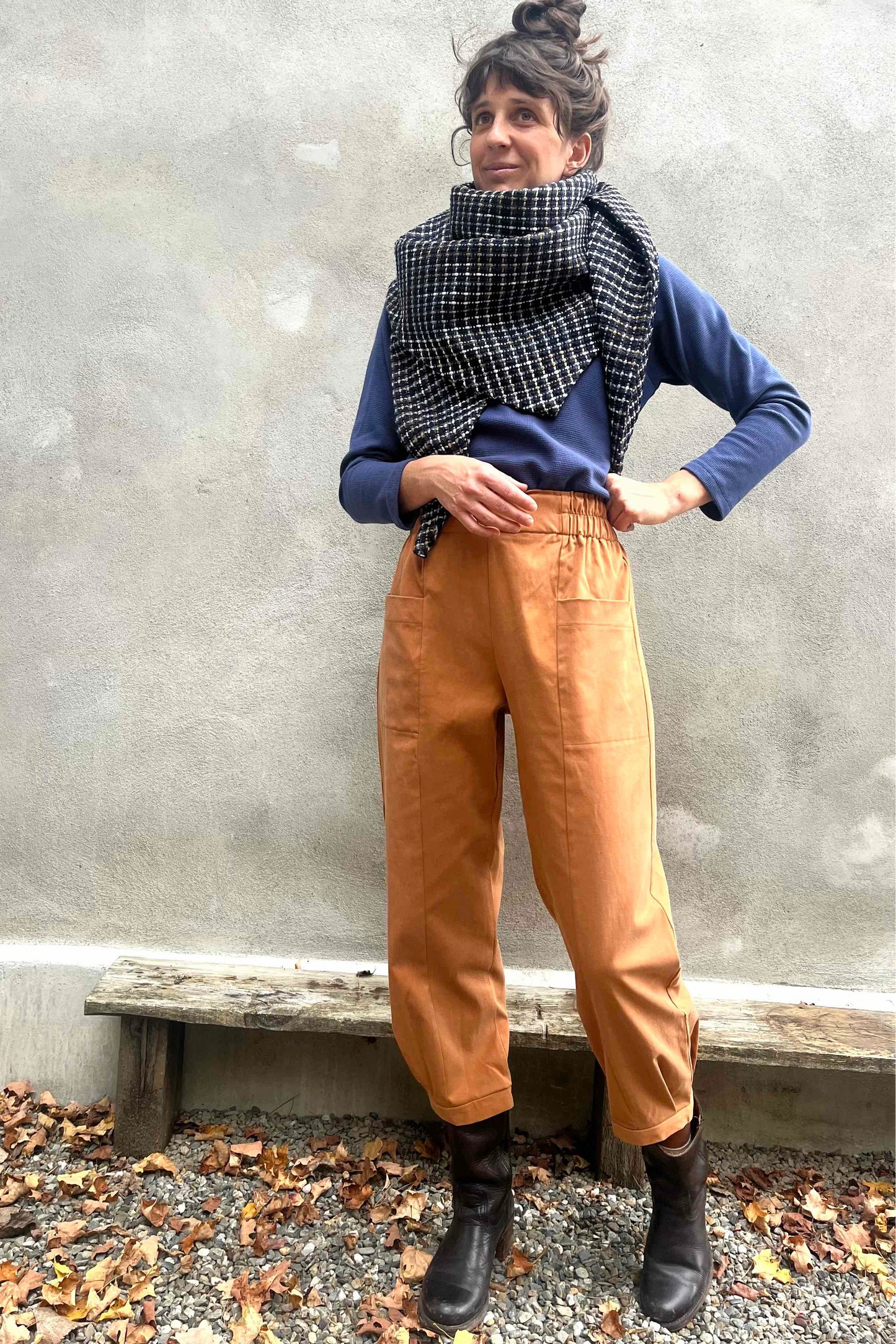 A woman wearing the Nyssa Pants by Kazak in Terracotta, barrel-legged pants with an elastic waistband and patch pockets. She is wearing them with the Oris Turtleneck in Indigo Waffle and standing outside by a grey wall. 