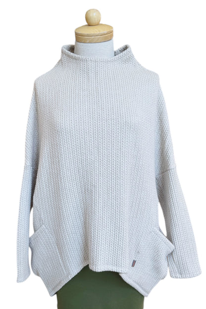 The Meeso Sweater by MOOVment in Oatmeal, a One Size oversized knit sweater with a wide mock turtleneck, long dropped sleeves and large side pockets, is shown on a mannequin in front of white background. 
