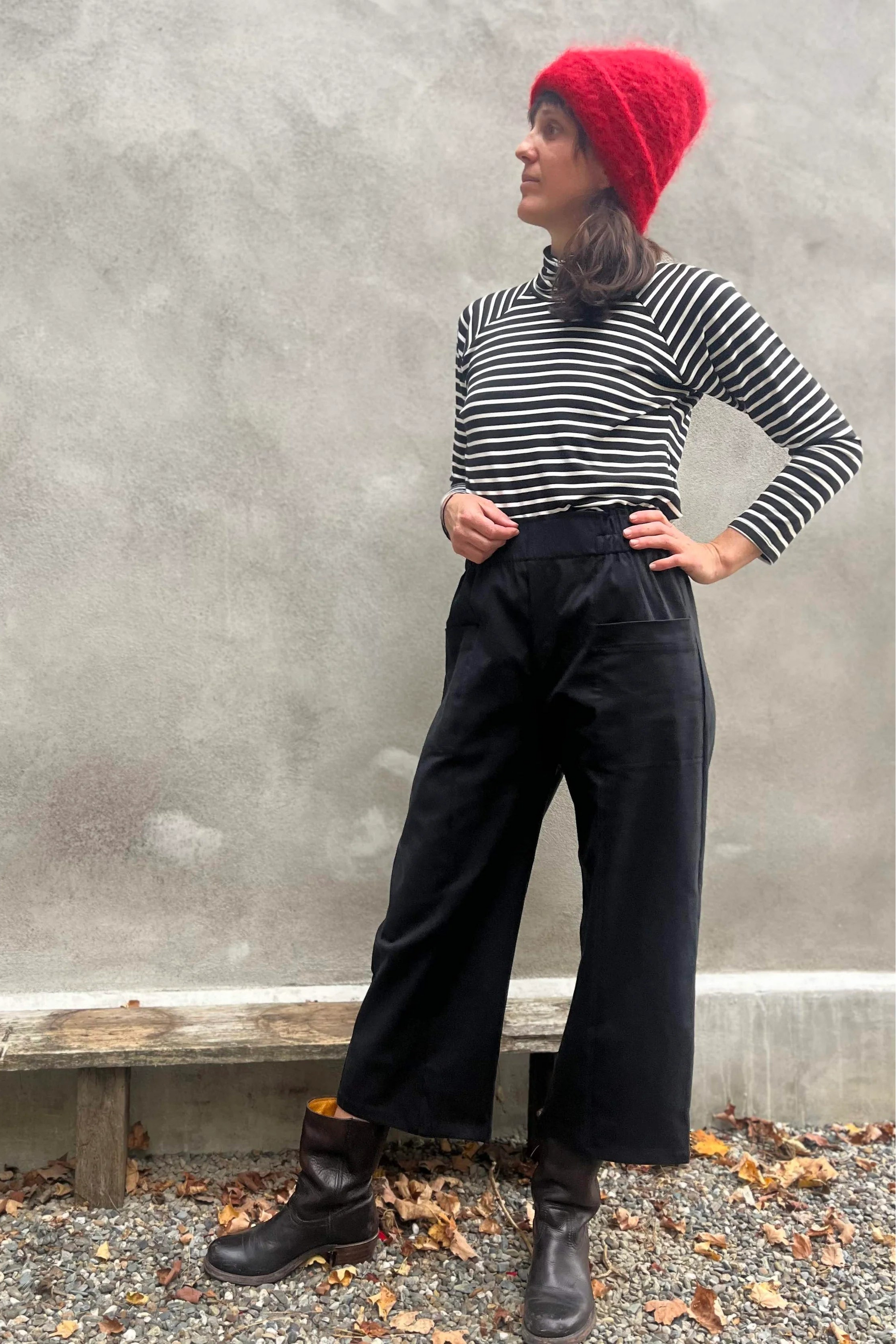 A woman wearing the Olea Pants by Kazak in Black, wide-legged ankle-length pants with a flat-front elastic waist and large patch pockets. She is wearing them with the Oris Turtleneck in Black Stripe and standing outside by a grey wall. 
