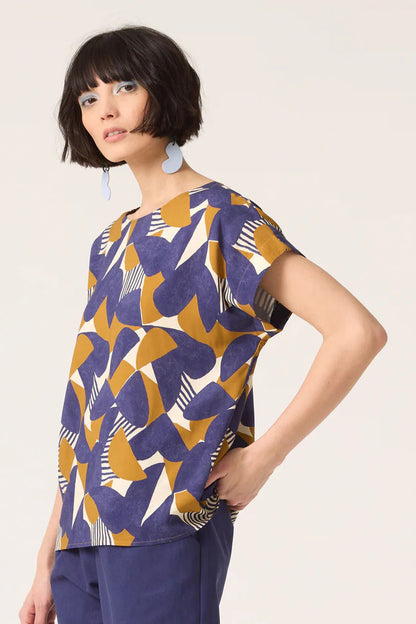 3/4 shot of a woman wearing the Ombre Top by Cokluch in Cumin Half Moon print featuring a round neck, short dropped sleeves, and a loose fit in OEKO-TEX certified viscose, with blue pants, standing in front of white background 