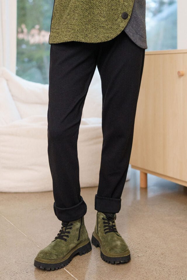 Waist down view of a woman wearing the Lichen Pant by Rien ne se Perd in Black, skinny leg pull-on pants in stretchy corduroy fabric. She is wearing them with a green and grey top and standing by a white chair. 