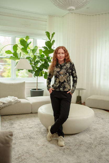 A woman wearing the Murphy Pants by  Rien ne se Perd, pull-on pants with a smooth front waistband, elastic at the back, and a slim fit with a bootcut hem. She is wearing them with black long-sleeve top with gold and white swirl patterns, and beige sneakers, standing in a modern living room with a round white ottoman.