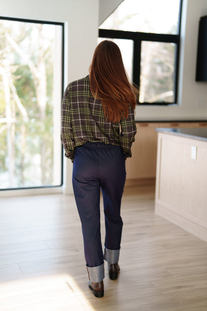 Back view of a woman wearing the Yamaska Pants by Rien ne se Perd in Denim Blue, pull on style jeans with rolled cuffs. She is wearing them with a green and brown plaid shirt, and dark brown ankle boots in a bright room with light wood flooring