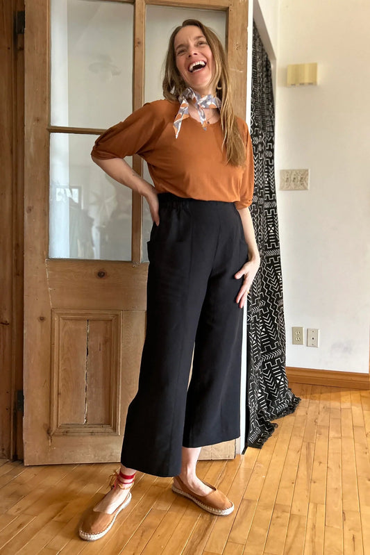A woman wearing the Petunia Pants by Kazak in Black, wide legged cropped pants with a high waist that's smooth in front with elastic at the back, and large patch pockets. She is wearing them with an orange top and standing in front of a wooden door. 