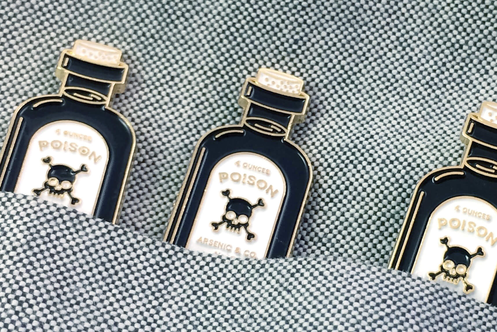 Three Poison Pins by Badaboöm Studio, featuring a tiny bottle of poison, are shown tucked into a textured blue pocket. 