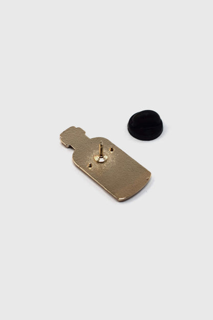 A back view of the Poison Pin by Badaboöm Studio, featuring a tiny bottle of poison, is shown against a white background. 