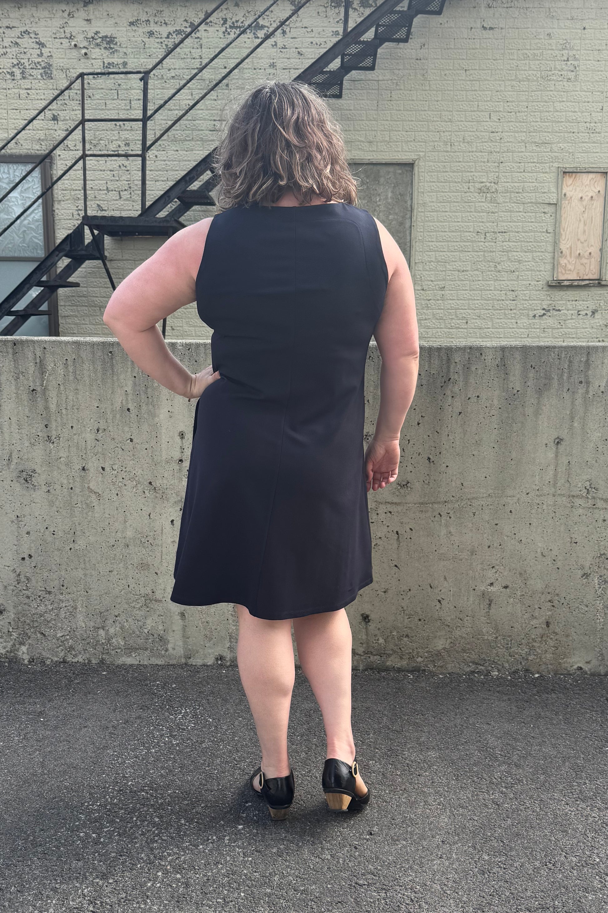 Back view of a woman wearing the Felicity Dress by Pure Essence in Navy, a sleeveless dress with a round neck, an a-line shape, and a knee length hem. She is standing in front of a brick building. 