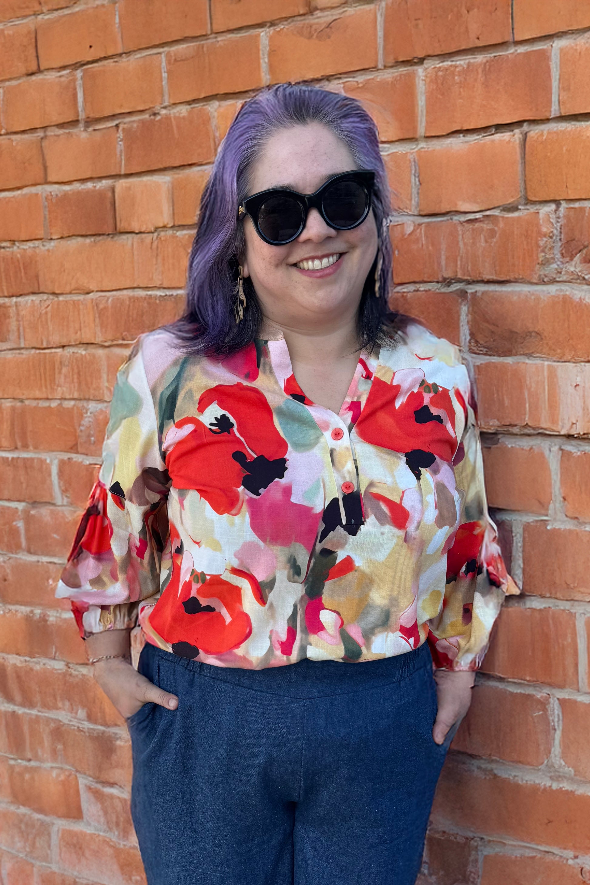 A woman wearing the Grace Blouse by Pure Essence in Coral Meadow, with a v-placket neck with buttons and 7/8 gathered sleeves. She is wearing it with denim pants and by a brick wall. 