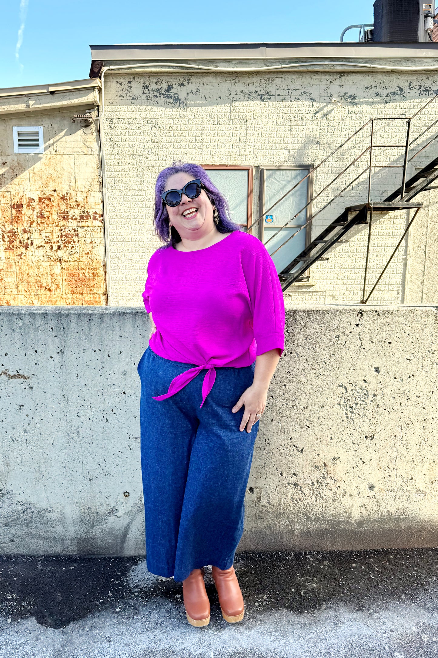 A woman wearing the Mateo Pants by Melow in Denim, ¾ wide leg pants with pull-on waistband that’s elasticized at the back. She is wearing them with a pink top and standing outside in front of a brick building. 