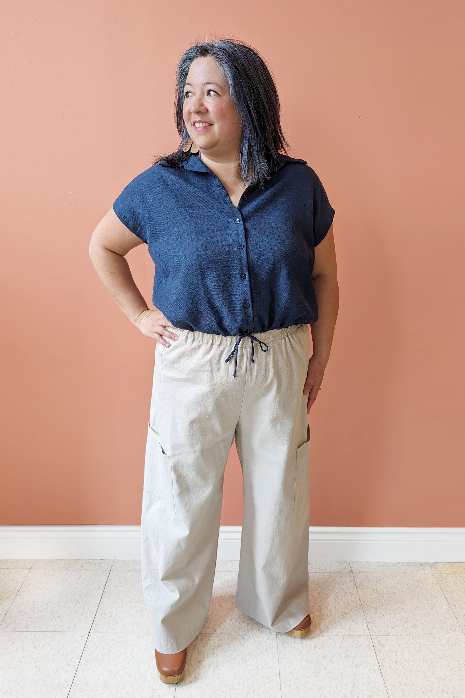 Christina is wearing the Chelsea Shirt by Pure Essence in Indigo with the Faro Pants by MAS in Oat, standing in front of a coral wall 