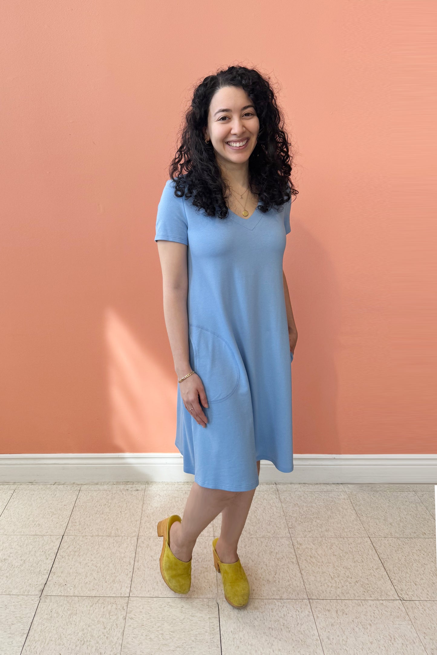 A woman wearing Gaia Dress by Pure Essence Oxford Blue, a short sleeved knee length dress with a V-neck, A-line shape, and pockets. She is standing against a coral wall.