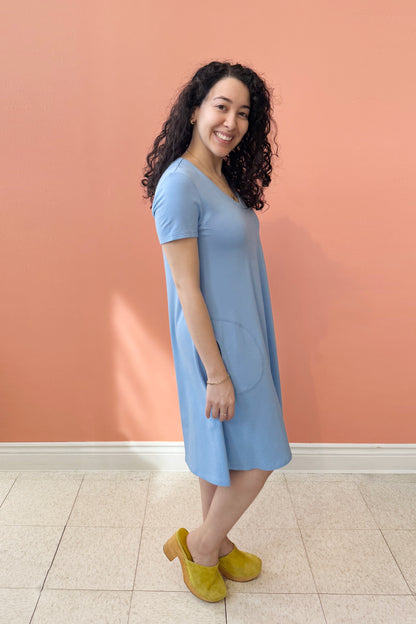 Side view of a woman wearing Gaia Dress by Pure Essence Oxford Blue, a short sleeved knee length dress with a V-neck, A-line shape, and pockets. She is standing against a coral wall.