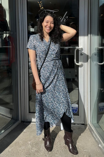A woman wearing the Galea Dress by Pure Essence in Blue Lattice print. This faux-wrap dress has short sleeves, a gathered waist with a tie and a midi-length tulip hem. She is standing outside the Flock entrance.