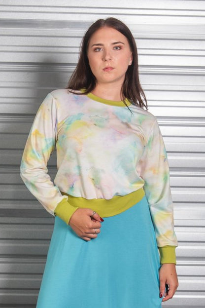 A woman wearing the Wanda E26 Sweater by Slak in Watercolour print, with a crew neck, long sleeves, and contrasting green ribbing at the cuffs, collar, and hem. She is wearing it with a a blue skirt and standing in front of a metallic shuttered door.