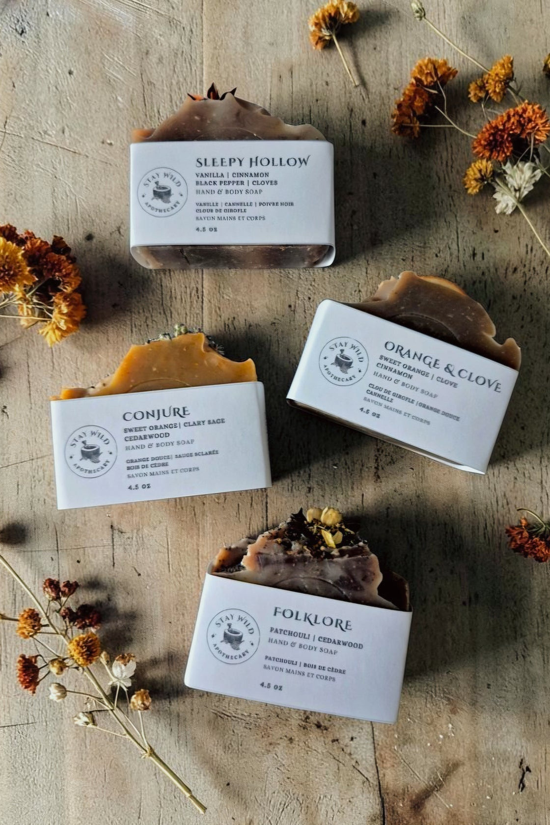 Four Stay Wild Apothecary bars of soap in white paper packaging, arranged on a rustic wooden surface with dried orange flowers. The soaps show different natural, earthy tones from the top: brown, golden yellow, and dark brown with botanical inclusions.