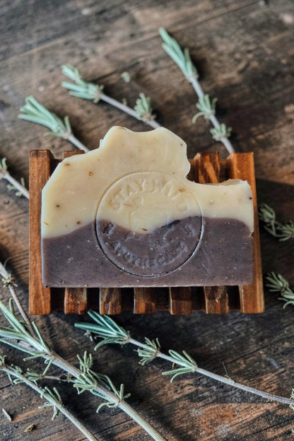 A handmade Witch's Garden soap bar from Stay Wild Apothecary, with a cream-coloured top half and a dark brown bottom half, displayed on a small wooden stand over a rustic dark wood surface with sprigs of rosemary.