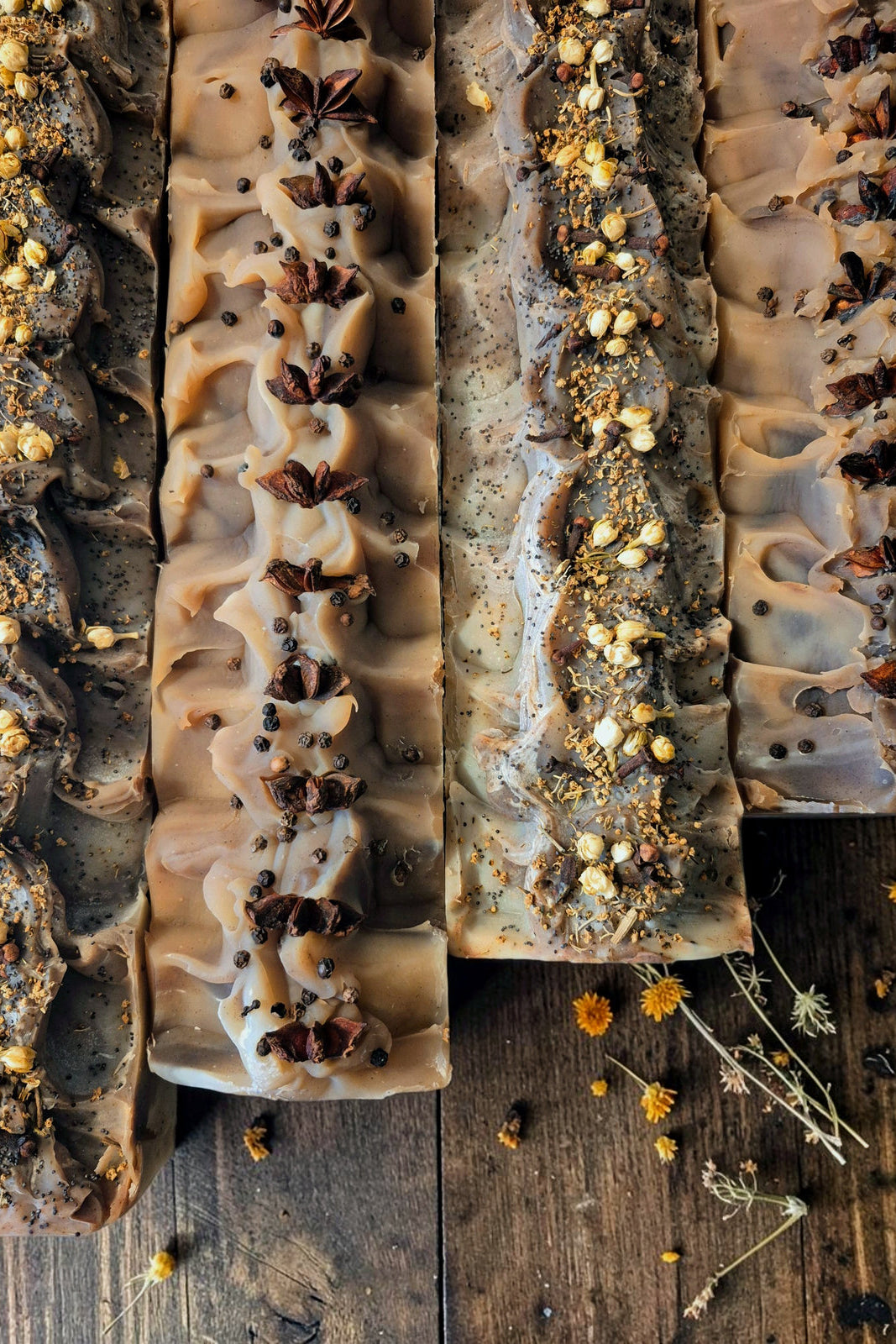 Four rectangular bars of Stay Wild Apothecary soap with textured ruffles, decorated with star anise, dried herbs, and seeds, arranged on a rustic dark wooden surface.