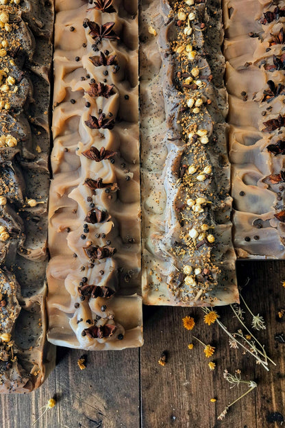 Four rectangular bars of Stay Wild Apothecary soap with textured ruffles, decorated with star anise, dried herbs, and seeds, arranged on a rustic dark wooden surface.