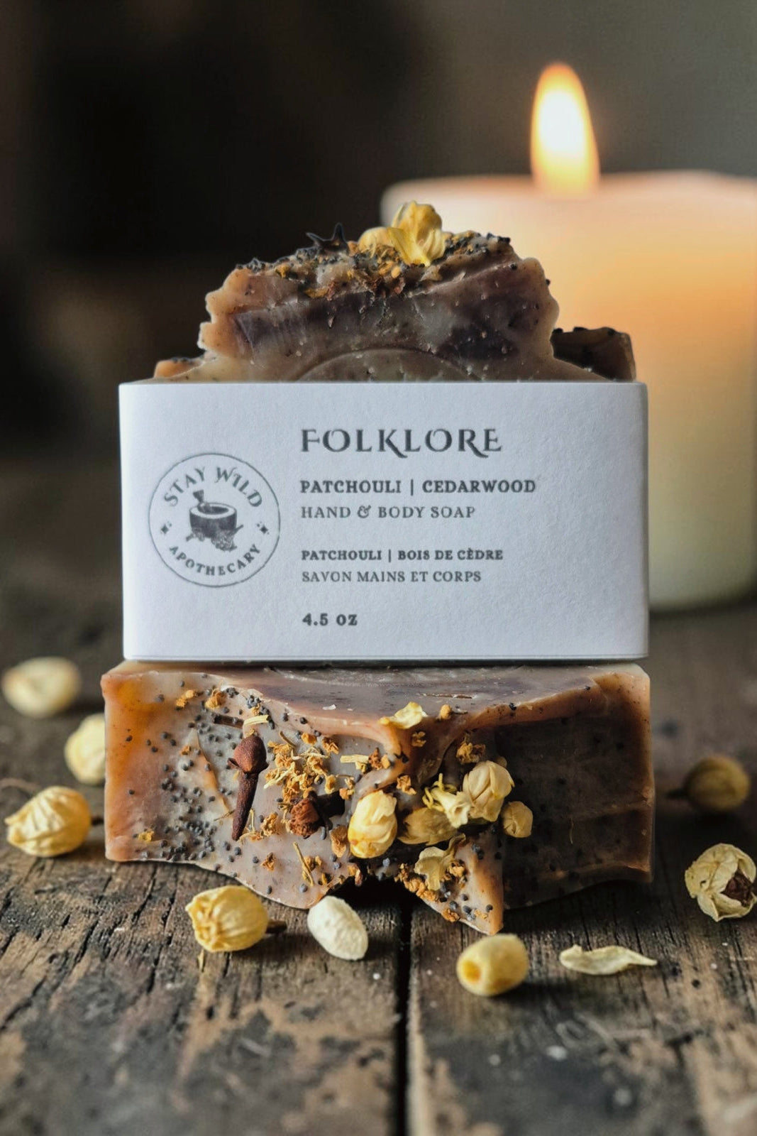 Two bars of Folklore Soap by Stay Wild Apothecary are stacked on a wooden table with a lit candle behind them. 