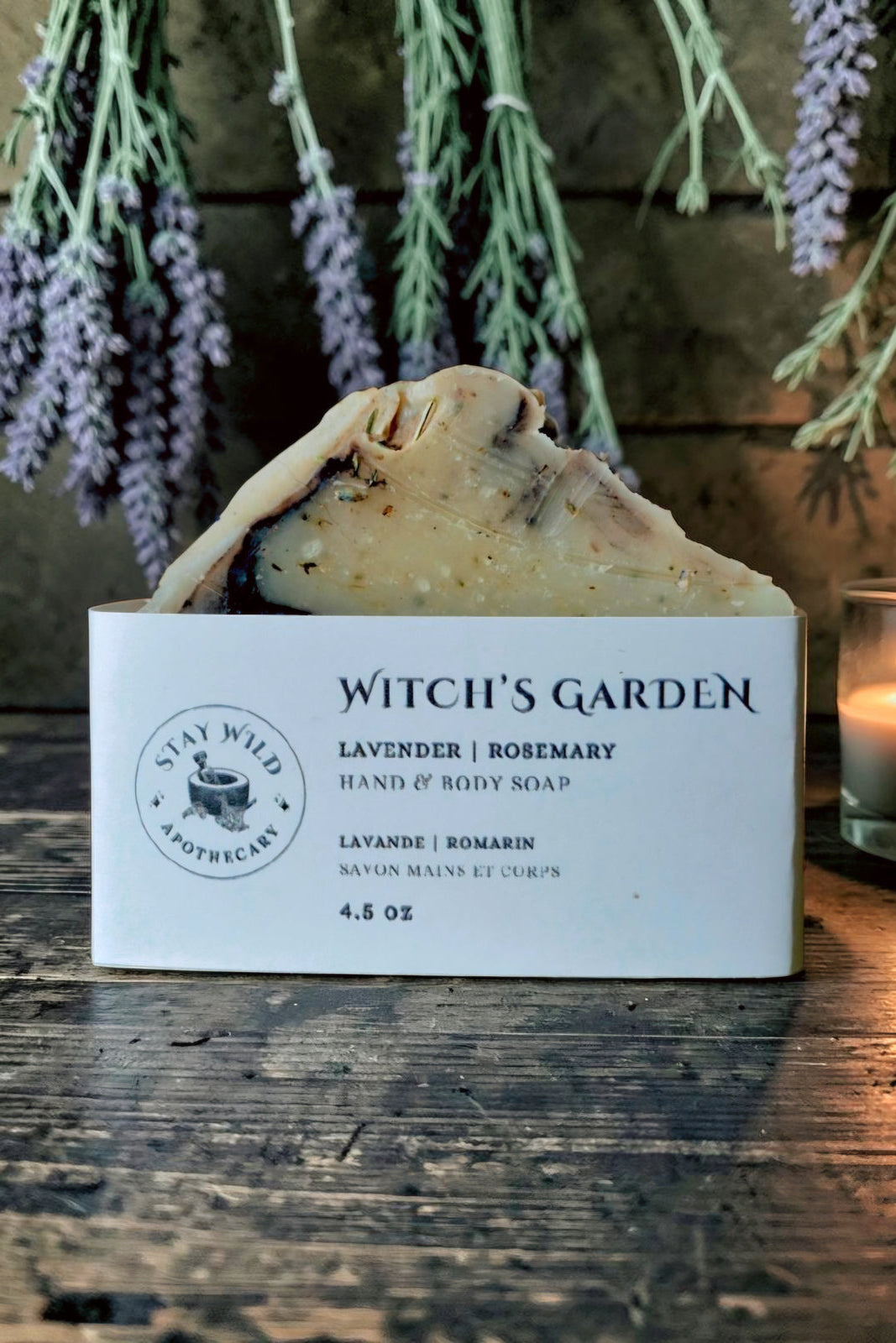 A handmade Witch's Garden soap bar from Stay Wild Apothecary, with a cream-coloured top half and a dark brown bottom half, displayed on wooden table with dried lavender hanging behind it