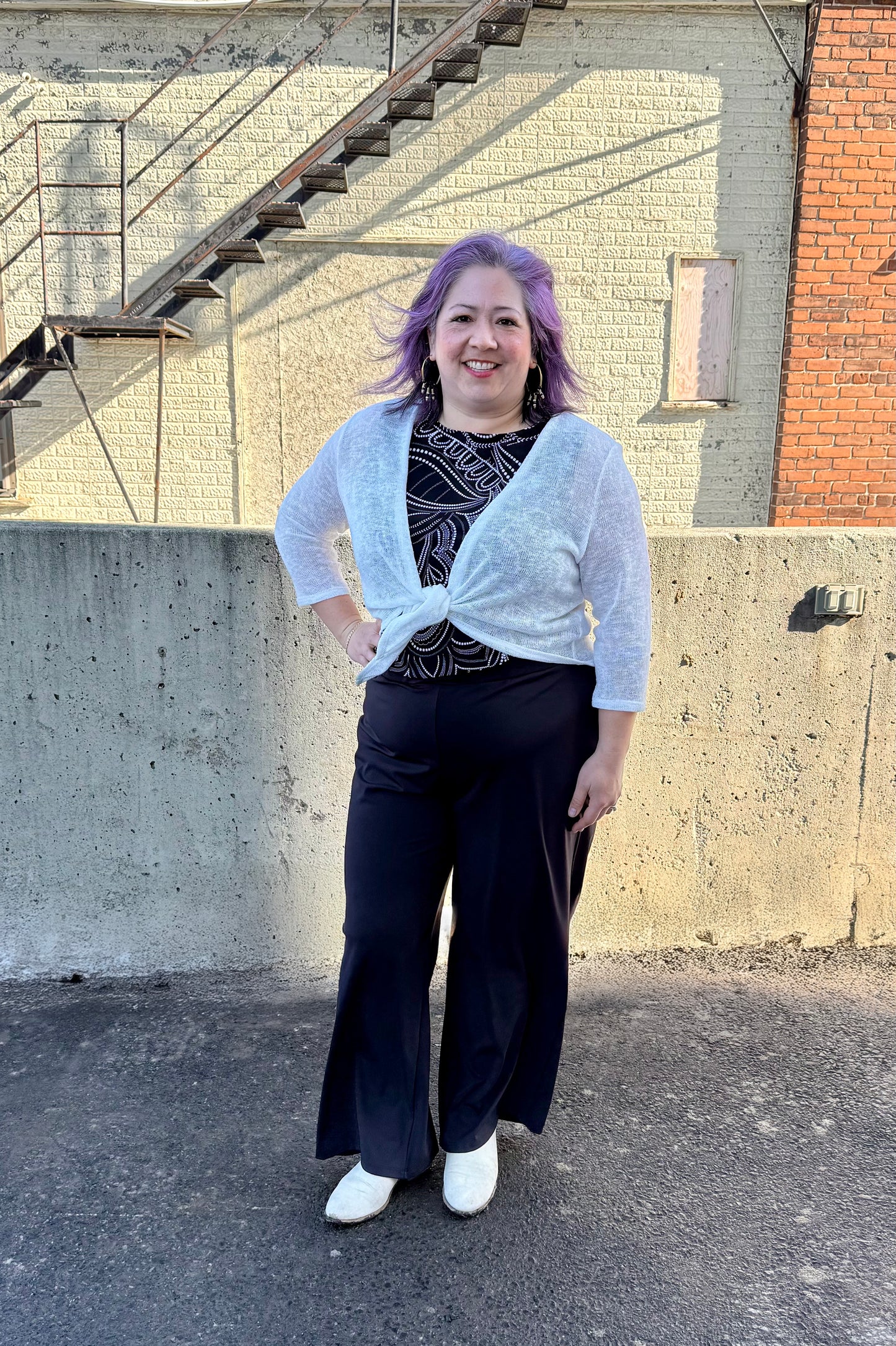 A woman wearing the Raina Tie-Front bolero by Compli K in Ivory.  She is wearing it with a black and white top and black pants and standing outside in front of a brick building. 