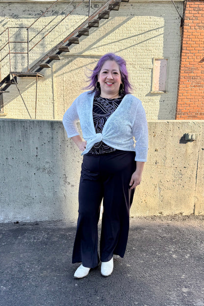 A woman wearing the Raina Tie-Front bolero by Compli K in Ivory.  She is wearing it with a black and white top and black pants and standing outside in front of a brick building. 