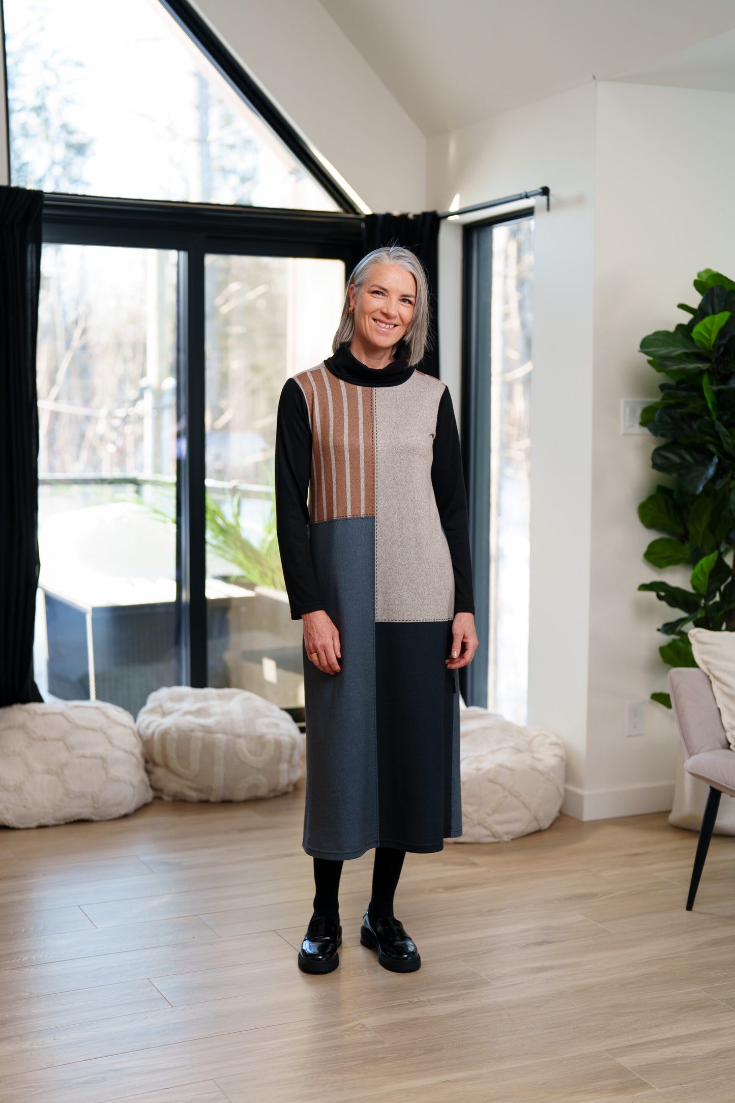 A woman wearing the Bonaventure Dress by Rien ne se Perd, a colour-blocked turtleneck sweater dress with panels of solid grey, blue and black, and brown stripes. She is standing in a room with bean bag chairs behind her. 