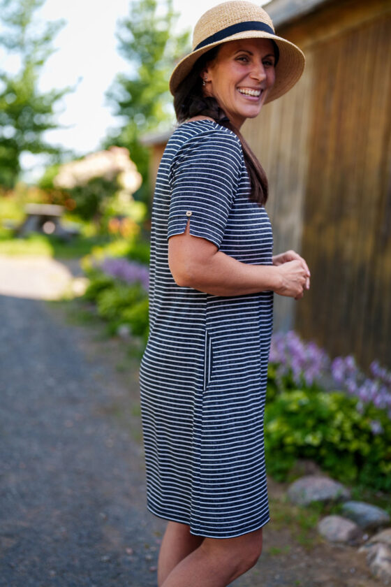 Side view of a woman wearing the Boogie Dress by Rien ne se Perd in Grey/White Stripe, a t-shirt dress with a wide neck, side pocket, and slit and coconut button detail on the short sleeves. She is standing outside. 