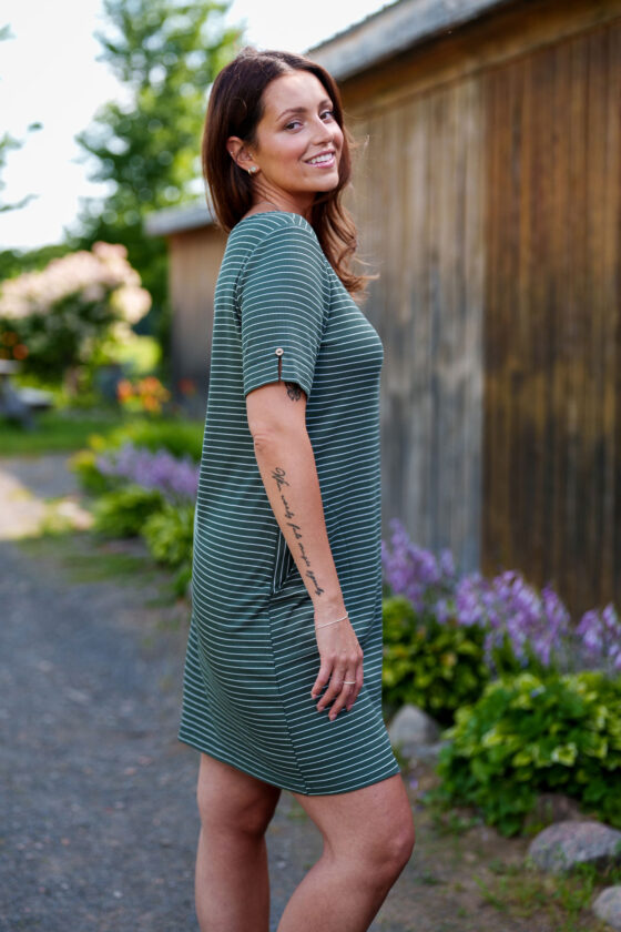 Side view of a woman wearing the Boogie Dress by Rien ne se Perd in Green/White Stripe, a t-shirt dress with a wide neck, side pocket, and slit and coconut button detail on the short sleeves. She is standing outside. 