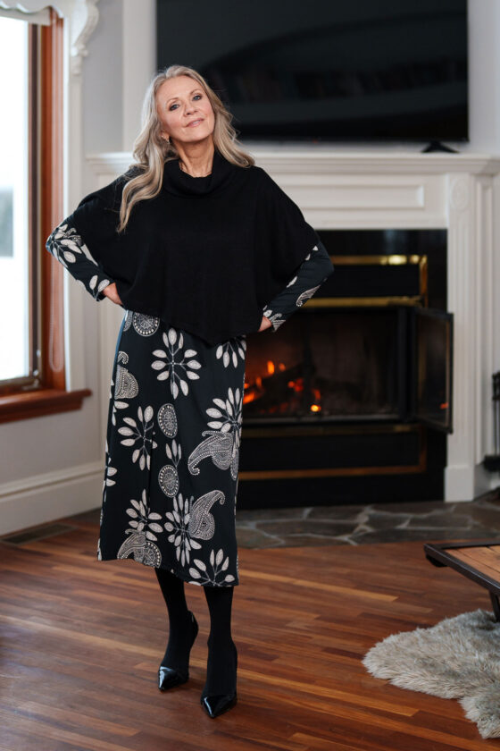 A woman wearing the Delacroix Bolero-Poncho by Rien ne se Perd in Black standing in front of fireplace