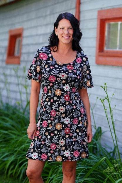 A woman wearing the Grenadine Dress by Rien ne se Perd in Floral print,  a short A-line dress with bell sleeves and a scoop neck. She is  standing in long grass in front of a rustic building with red windows.