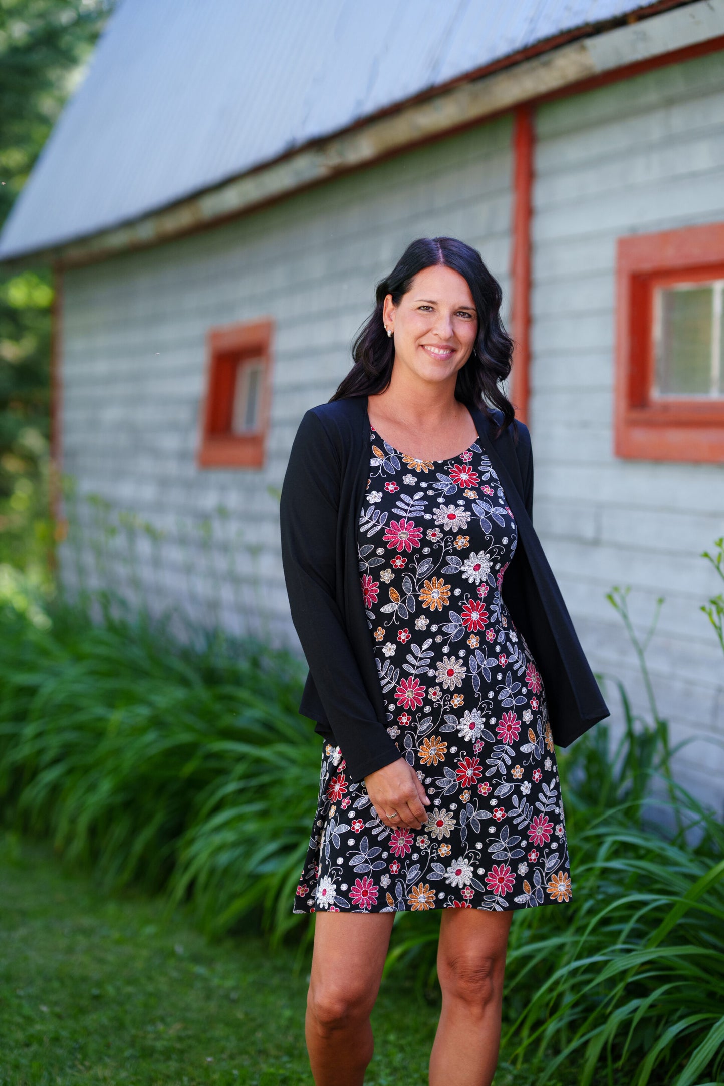 A woman wearing the Grenadine Dress by Rien ne se Perd in Floral print,  a short A-line dress with bell sleeves and a scoop neck. She is  wearing it under a black cardigan and standing in front of a rustic building with red windows.