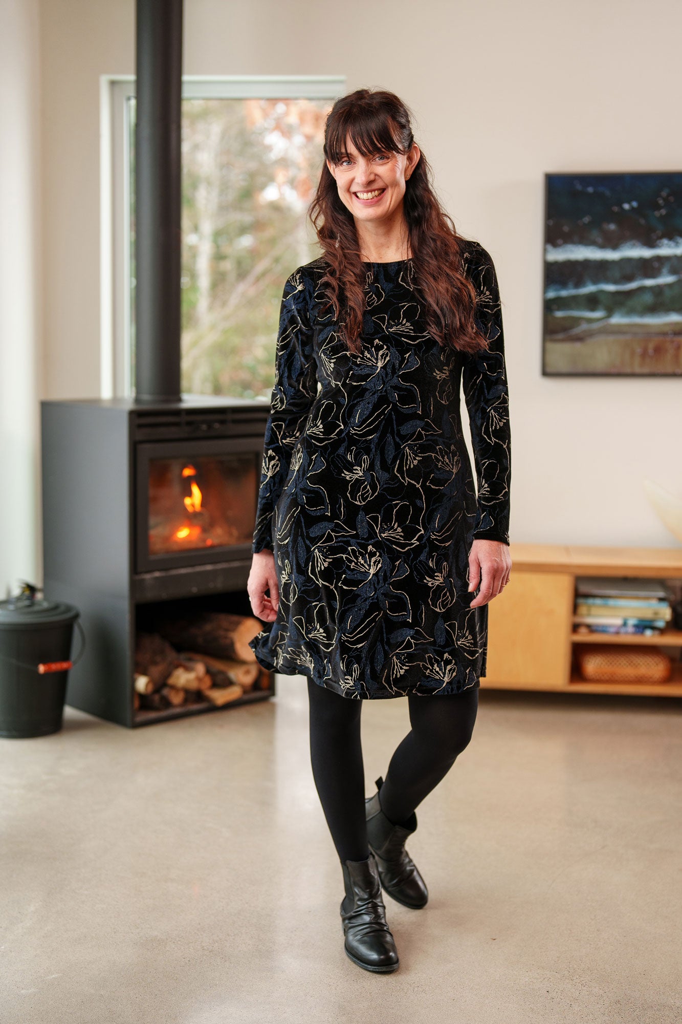 A woman wearing the Larix Dress by Rien ne se Perd in Black/Navy/Gold, a velvet dress with a floral pattern, a round neck, an a knee-length A-line shape. She is standing in front of a fireplace.