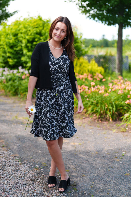 A woman wearing the Mambo Dress from Rien ne se Perd, a black and white floral dress with a V-neck, short sleeves, adjustable empire waist, and knee-length A-line skirt. She is standing outside in a garden. 