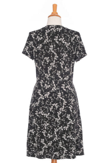 A back view of the Mambo Dress from Rien ne se Perd, a black and white floral dress with a V-neck, short sleeves, adjustable empire waist, and knee-length A-line skirt, is shown on a mannequin in front of a white background. 