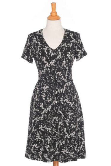 The Mambo Dress from Rien ne se Perd, a black and white floral dress with a V-neck, short sleeves, adjustable empire waist, and knee-length A-line skirt, is shown on a mannequin in front of a white background. 