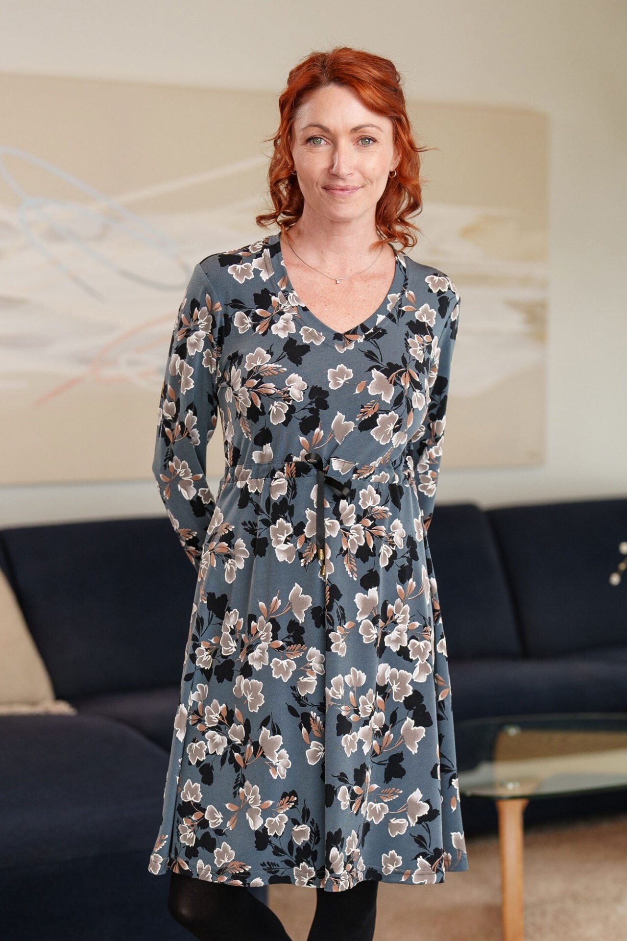 A woman wearing the Mariana Dress by Rien ne se Perd in Blue Floral, featuring a V-neck, an empire waist with a ribbon detail, long sleeves, and a knee-length A-line skirt. She is standing in a living room setting.