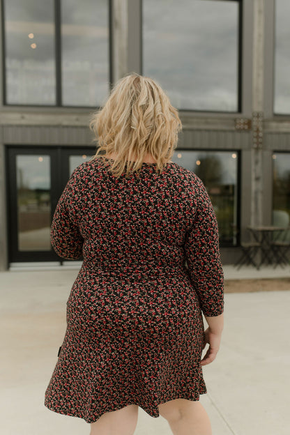 Back view of a woman wearing the Mattawin Dress from Rien ne se Perd in Red Floral, an above-the-knee length faux-wrap dress with three-quarter sleeves. She is standing outside a concrete building. 