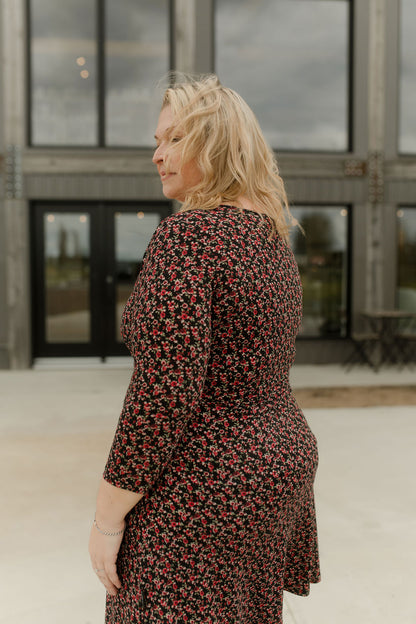 Side view of A woman wearing the Mattawin Dress from Rien ne se Perd in Red Floral, an above-the-knee length faux-wrap dress with three-quarter sleeves. She is standing outside a concrete building. 