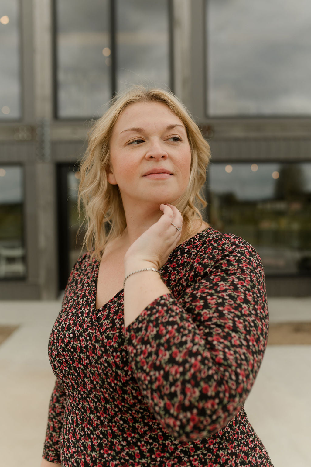 Close up of a woman wearing the Mattawin Dress from Rien ne se Perd in Red Floral, an above-the-knee length faux-wrap dress with three-quarter sleeves. She is standing outside a concrete building. 