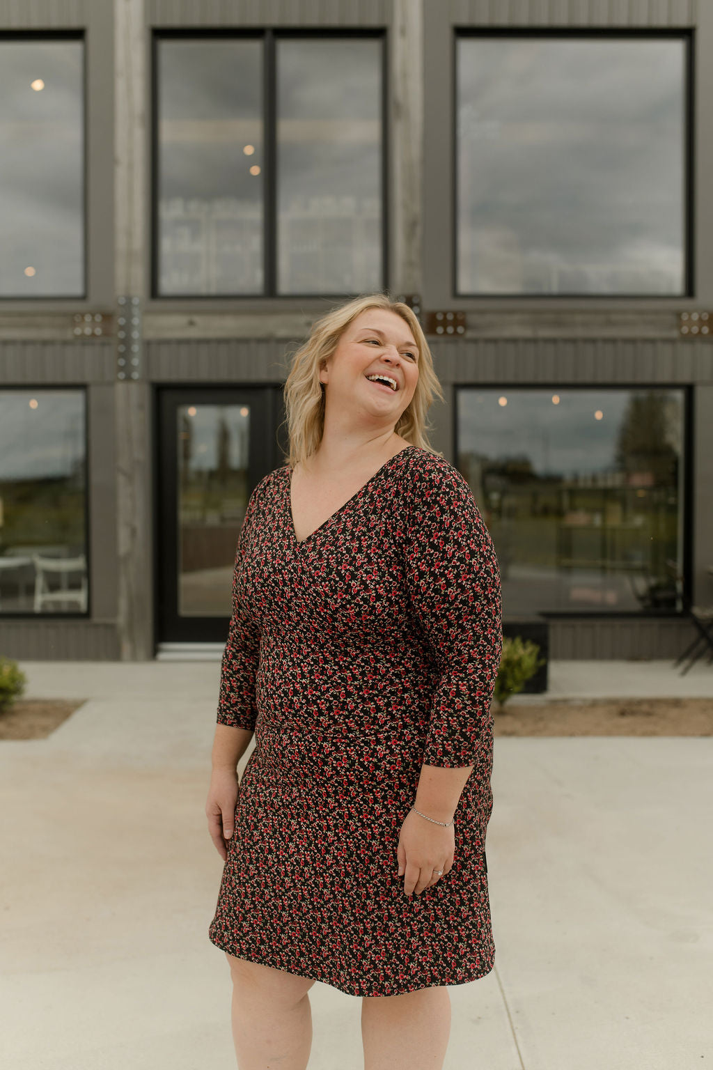 A woman wearing the Mattawin Dress from Rien ne se Perd in Red Floral, an above-the-knee length faux-wrap dress with three-quarter sleeves. She is standing outside a concrete building. 