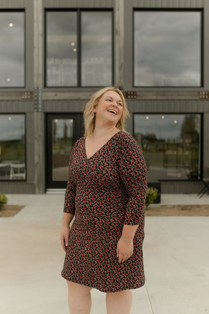 A woman wearing the Mattawin Dress from Rien ne se Perd in Red Floral, an above-the-knee length faux-wrap dress with three-quarter sleeves. She is standing outside a concrete building. 