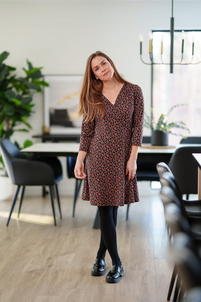 A woman wearing the Mattawin Dress from Rien ne se Perd in Red Floral, an above-the-knee length faux-wrap dress with three-quarter sleeves. She is standing in a modern, softly blurred dining room setting.
