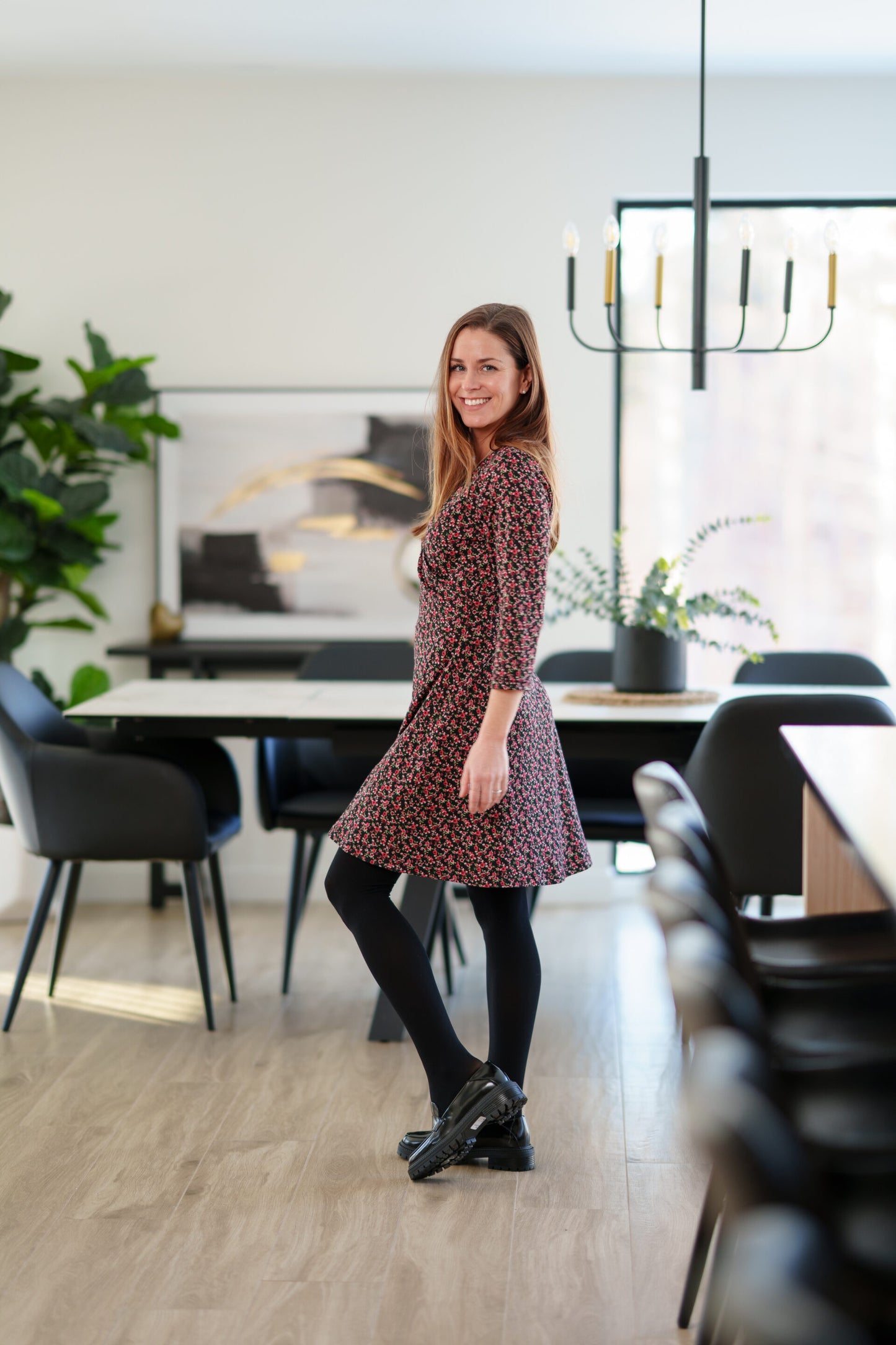 Side view of a woman wearing the Mattawin Dress from Rien ne se Perd in Red Floral, an above-the-knee length faux-wrap dress with three-quarter sleeves. She is standing in a modern, softly blurred dining room setting.
