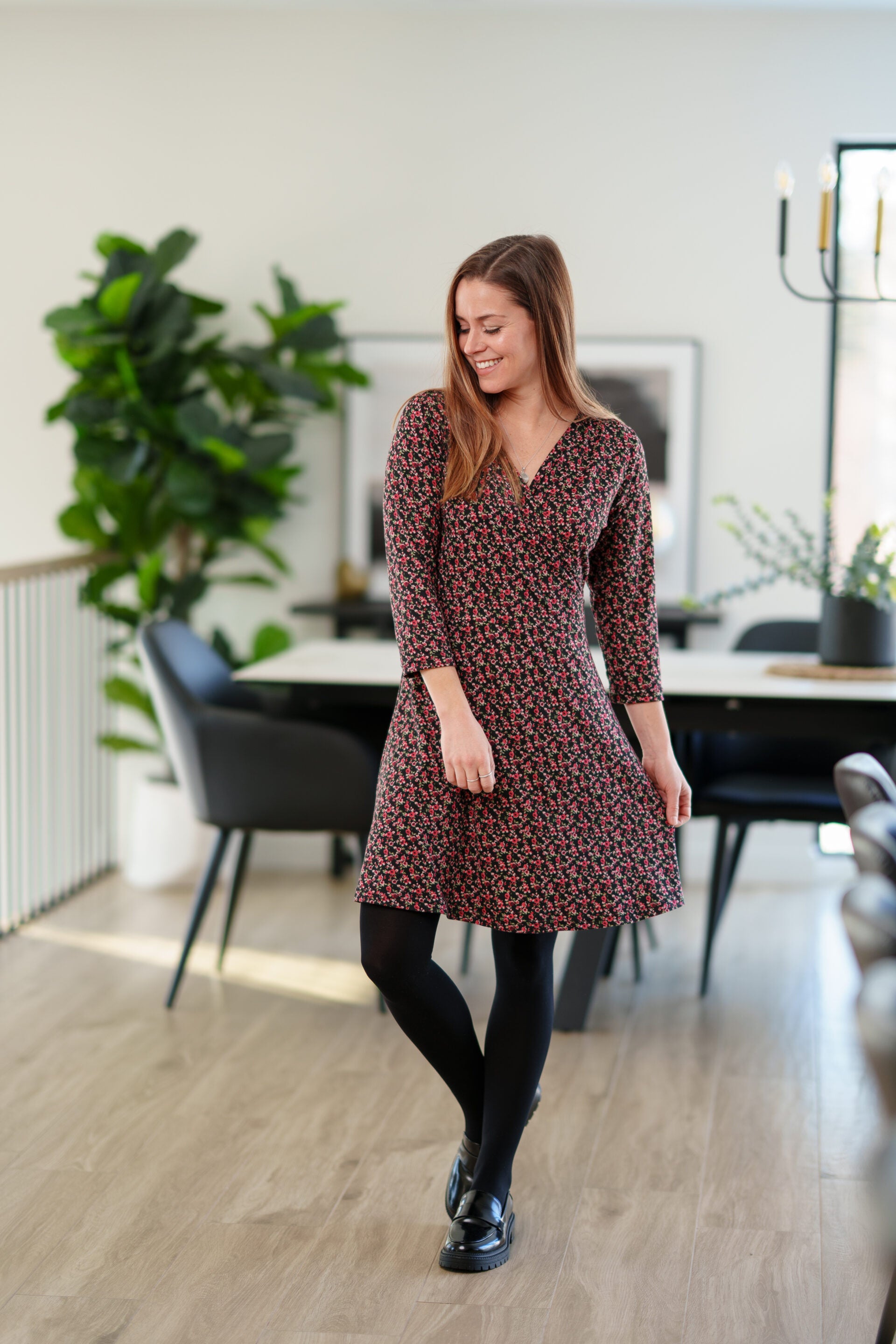 A woman wearing the Mattawin Dress from Rien ne se Perd in Red Floral, an above-the-knee length faux-wrap dress with three-quarter sleeves. She is standing in a modern, softly blurred dining room setting.
