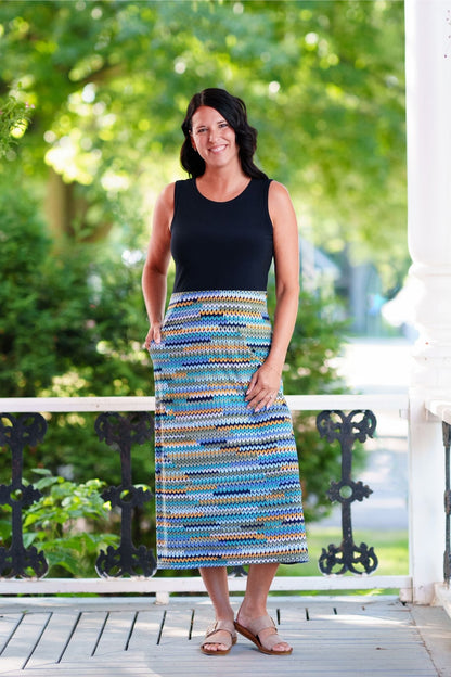 A woman wearing the Mimosa Dress by Rien ne se Perd in Navy/Mesh print, a sleeveless 7/8 length dress with a solid navy top and a straight skirt with a mesh print in blue tones. She is standing on a wooden porch. 