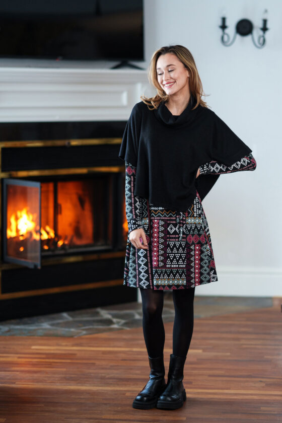 A woman wearing the Delacroix Bolero-Poncho by Rien ne se Perd in Black standing in front of fireplace